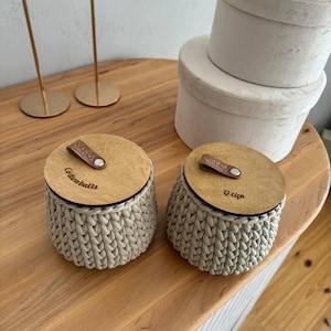Puede incluir: Dos contenedores de almacenamiento de crochet beige con tapas de madera. Las tapas tienen correas de cuero y est&aacute;n etiquetadas como "Cottonballs" y "Q-tips".