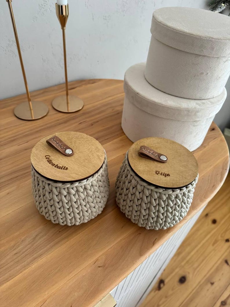 Puede incluir: Dos contenedores de almacenamiento de crochet beige con tapas de madera. Las tapas tienen correas de cuero y est&aacute;n etiquetadas como "Cottonballs" y "Q-tips".