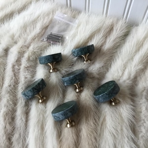 May include: Six cabinet knobs with a round, dark green marble top and a gold-colored base. The knobs are arranged on a white fur-like surface, with a small bag of screws visible in the background. These knobs are ideal for furniture upgrades.