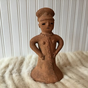May include: A terracotta figurine of a person with arms akimbo, wearing a hat and a dress. The figure has a necklace and a zig-zag pattern down the front. The statue is on a white fur-like surface.
