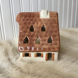 May include: A ceramic house-shaped candle holder with a brown roof and white walls. The roof has several triangular cutouts and a small chimney. The house sits on a fluffy, white surface.