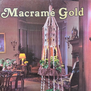 Peut inclure: Un porte-plante en macramé à deux niveaux, dans des tons bruns et beiges. Le porte-plante est suspendu au plafond et contient deux plantes en pot. L'image est tirée d'un livre intitulé "Macramé Gold" de Jeep Ziemer & Karol Smith.
