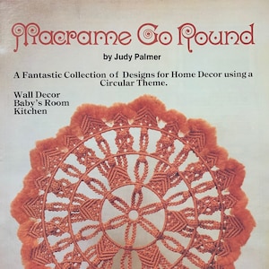 May include: A close-up of a large, round macrame wall hanging with a circular design. The macrame is made with a thick, orange cord and has a fringe border. The text "#7430, Plaid" is visible in the lower right corner of the image.