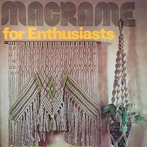 May include: A book cover with the title "Macrame for Enthusiasts" in bold, black letters. The cover features a macrame wall hanging with a geometric pattern and a macrame plant hanger with a green plant. The text "Easy to follow Knotting Diagrams" and "Instructions for 12 projects including wall hangings, pot hangers, macrame jewelry." is also visible on the cover.