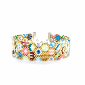Puede incluir: Un brazalete de cuentas coloridas con un patrón geométrico en tonos de azul, verde, rosa y dorado. El brazalete está hecho con cuentas pequeñas y redondas y tiene un cierre plateado.