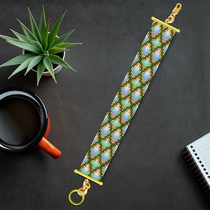 Może przedstawiać: Bransoletka z koralików z geometrycznym wzorem w odcieniach różowego, zielonego i żółtego. Bransoletka ma złote zapięcia na obu końcach.