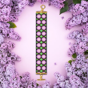 May include: A beaded bracelet featuring a geometric design of pink, green and white flowers on a black background. The bracelet has gold-toned clasps.