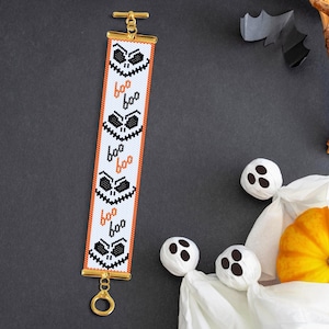 May include: A white beaded bracelet with a repeating pattern of black and orange Halloween-themed faces. The bracelet has a gold clasp and a gold toggle.