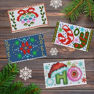 May include: Four beaded Christmas cards with festive designs. One card features a red bow and ornament, another has a snowflake design, a third says "JOY" with a candy cane, and the last says "HO HO HO" with a Santa hat and ornament. The cards are on a wooden surface.