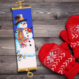 May include: A beaded bracelet featuring a snowman design. The snowman is wearing a blue hat, a colourful scarf, and a red flower. The bracelet is made with white and blue beads and has a gold clasp.