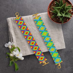 以下が含まれることがあります： 金色の留め具が付いたカラフルなビーズのブレスレット2本。ブレスレットは、赤、オレンジ、黄色、緑、青、紫の色合いの幾何学模様が特徴です。