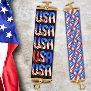 May include: Two beaded bracelets with a patriotic theme. One bracelet features the word "USA" repeated five times in red, white, and blue beads on a black background. The other bracelet features a repeating pattern of red, white, and blue stars and stripes.