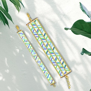 May include: Two beaded bracelets with a geometric pattern in shades of blue, green, and white. The bracelets have gold-colored clasps and end caps. The bracelets are displayed on a white surface with shadows and greenery.