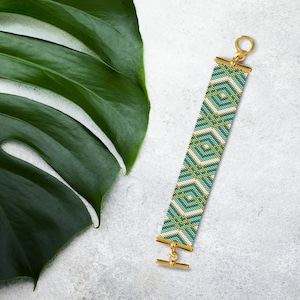 May include: A white, blue, and gold beaded bracelet with a geometric pattern. The bracelet has a gold clasp and is lying on a white surface with a green leaf.