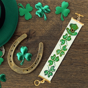 May include: A white beaded bracelet with a green shamrock pattern and a gold clasp. The bracelet features a small green leprechaun hat charm. The bracelet is surrounded by green shamrocks and a horseshoe.