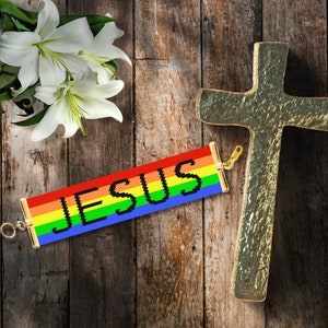 May include: A rainbow-colored beaded bracelet with the word "JESUS" spelled out in black beads. The bracelet is on a wooden background with a gold cross.