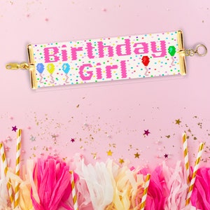 May include: A pink and white beaded bracelet with the words "Birthday Girl" and colourful balloons. The bracelet is on a pink background with gold confetti.