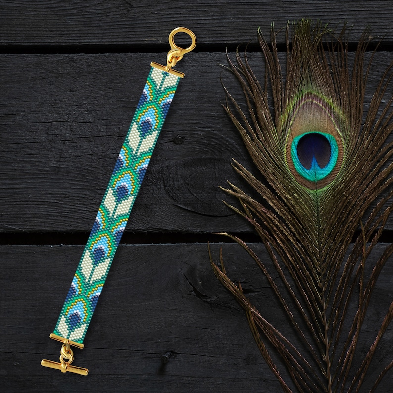 May include: A beaded bracelet with a peacock feather design in shades of blue, green, and white. The bracelet has a gold clasp and is on a black wooden surface.