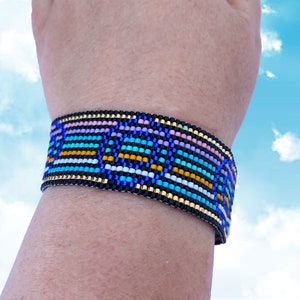 May include: A colorful beaded bracelet with a geometric pattern in shades of blue, pink, yellow, white, and black. The bracelet is worn on a person's wrist.