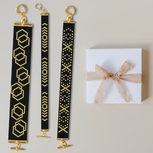 May include: Three black beaded bracelets with gold accents. The bracelets feature different geometric patterns. The bracelets are displayed on a light beige surface.
