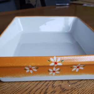 May include: A rectangular ceramic baking dish with a white interior and a vibrant orange exterior rim. The orange border is decorated with small, hand-painted white daisy flowers featuring red centres, arranged in a repeating pattern along the side.