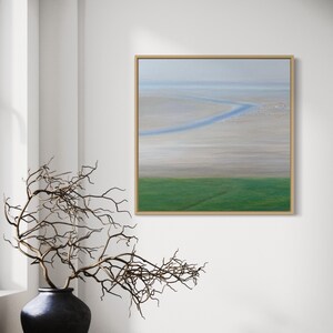 Könnte beinhalten: Gerahmtes Landschaftsgemälde mit einer ruhigen Küstenlandschaft. Das Kunstwerk zeigt ein grünes Feld, einen Sandstrand und einen blauen Wasserweg unter einem dunstigen Himmel. Eine dunkle Vase mit Zweigen steht auf einem Holztisch.