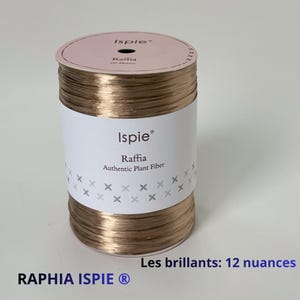以下が含まれることがあります： メタリックブロンズ色のIspieブランドのラフィア（天然植物繊維）の円筒形スプール。白い紙のラベルには「Ispie」「Raffia」「Authentic Plant Fiber」という文字と、小さな灰色の十字模様が印刷されています。上部は淡いピンク色です。