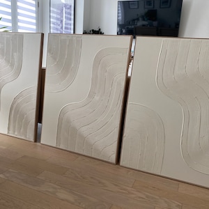 May include: Three white canvas wall art pieces with a raised, textured design of curved lines. The art pieces are framed in a light wood frame.