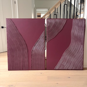 May include: A pair of rectangular wall art panels in a deep burgundy color. Each panel features a smooth, flat surface contrasted by sections of raised, vertical, and curved line textures that create a rhythmic, flowing pattern across the canvas.