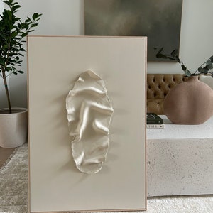 May include: A white canvas with a raised, textured, oval-shaped design in the center. The canvas is framed in a light wood frame.