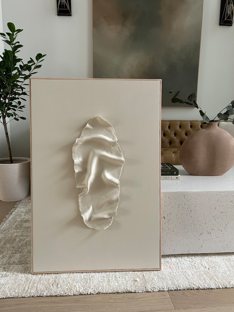 May include: A white canvas with a raised, textured, oval-shaped design in the center. The canvas is framed in a light wood frame.