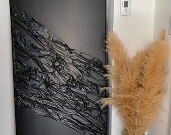 May include: A black framed canvas with a textured black plastic sheet draped over it. The plastic sheet is crumpled and has a diagonal pattern. The canvas is leaning against a white wall next to a vase with dried pampas grass.
