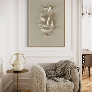 May include: A beige armchair with a knitted throw blanket, a white ceramic vase with two handles, and a gold side table with a marble base. A framed abstract art print with a white, textured, organic shape hangs on the wall behind the chair.