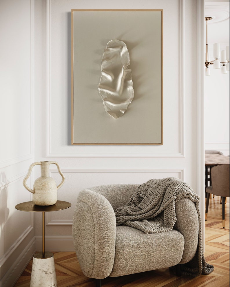 May include: A beige armchair with a knitted throw blanket, a white ceramic vase with two handles, and a gold side table with a marble base. A framed abstract art print with a white, textured, organic shape hangs on the wall behind the chair.