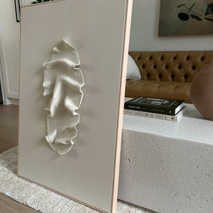 May include: A white, textured, 3D sculpture of a face on a beige canvas with a wooden frame. The sculpture is mounted on a white wall.