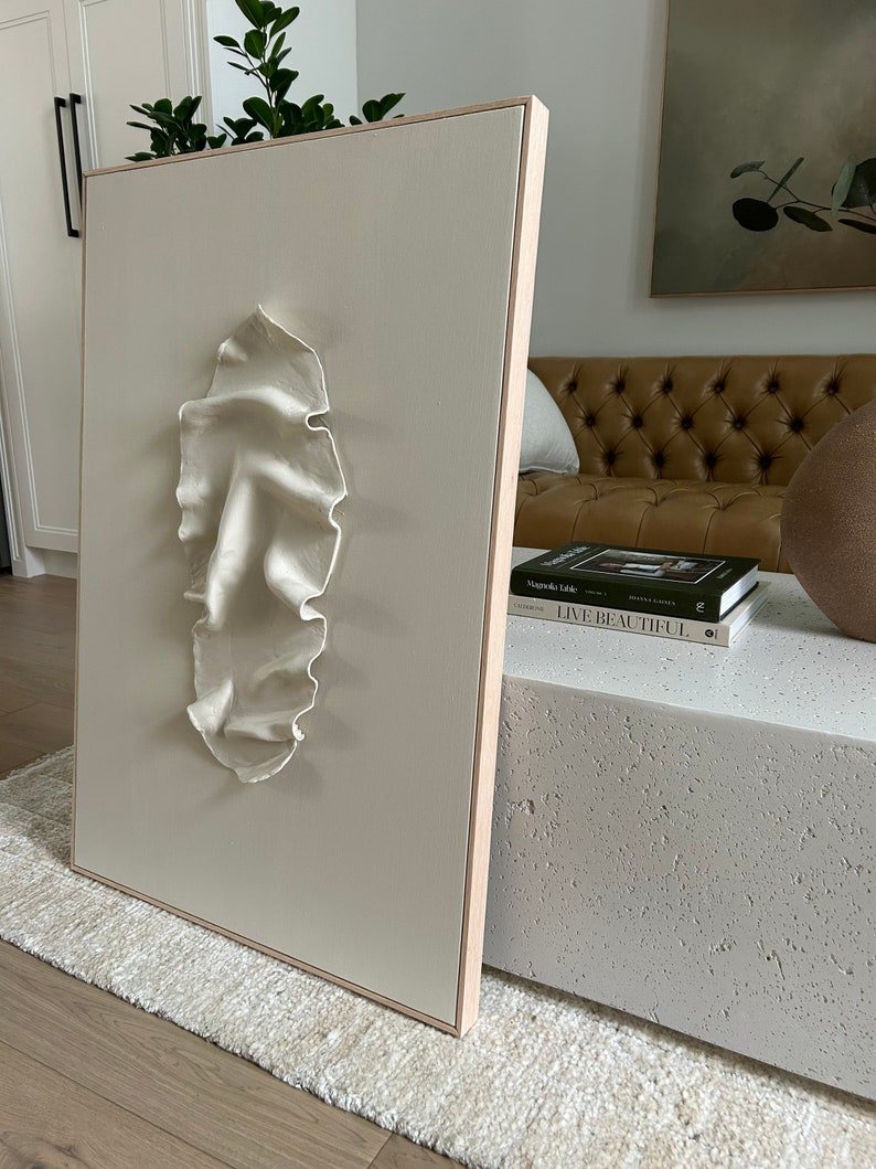 May include: A white, textured, 3D sculpture of a face on a beige canvas with a wooden frame. The sculpture is mounted on a white wall.
