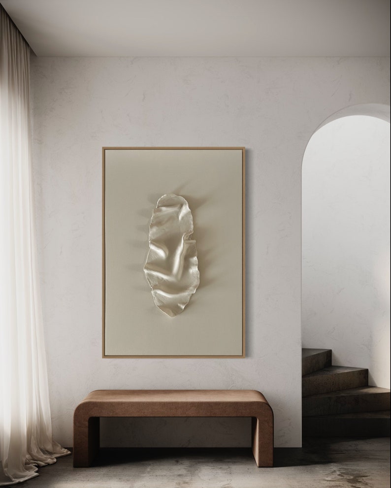 May include: A framed abstract art print with a white, textured, organic shape on a beige background. The print is hanging on a white wall above a brown wooden bench.