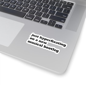 May include: A white sticker with the text "just hyperfixating on a new musical bootleg" in black, placed on a silver laptop. The laptop keyboard is visible in the background.
