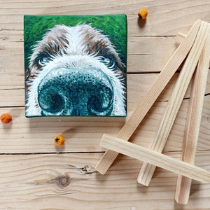 Puede incluir: Una pintura de primer plano de la cara de un perro en un lienzo pequeño. El perro tiene pelo marrón y blanco y una nariz grande y negra. La pintura está en un caballete de madera.