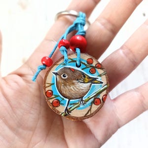 Op de afbeelding: Een handgeschilderde houten sleutelhanger met een klein vogeltje dat op een tak zit met rode bessen. De sleutelhanger is versierd met blauwe en witte verf en heeft een turquoise koord met rode kralen.