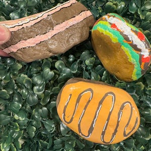 May include: Three hand-painted rocks shaped like food items. One rock resembles a brown layered cake with pink frosting and sprinkles. Another is painted to look like a burger with green lettuce and red accents. The third is a yellow rock with brown icing.