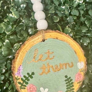 May include: A round wooden ornament with a light green center and a gold-colored border. The words "let them" are written in gold script. The ornament is decorated with painted flowers and greenery. A string of white beads is attached for hanging.