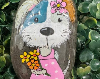 May include: A smooth, oval-shaped grey river rock hand-painted with a cartoon dog. The dog has white fur, a blue patch over one eye, and a pink flower on its head. It wears a pink dress with white polka dots and holds a bouquet of yellow flowers.