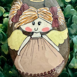 Puede incluir: Una piedra de río ovalada y lisa pintada a mano con una figura de ángel de dibujos animados. El ángel tiene cabello rubio en coletas con lazos rojo oscuro, un halo dorado, alas amarillas y un vestido color canela con detalles.