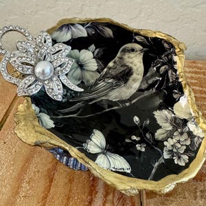 May include: A decorative oyster shell dish featuring a black interior with a white bird, floral patterns, and a butterfly. The shell edge is painted gold, and a silver-toned brooch with clear rhinestones and a central white pearl is attached.