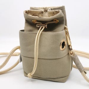 Könnte beinhalten: Eine kleine, olivgrüne Canvas-Bucket-Bag mit Kordelzugverschluss und einem langen, cremefarbenen Seilriemen. Die Tasche verfügt über goldfarbene Hardware, einschließlich Ösen und einem Reißverschluss. Das Innenfutter hat ein Muster.
