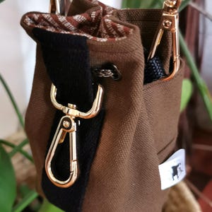 Puede incluir: Una bolsa de lona marr&oacute;n con una franja negra y herrajes dorados. La bolsa tiene un cierre de cord&oacute;n y un interior estampado. Se ve una peque&ntilde;a etiqueta blanca con una silueta negra.