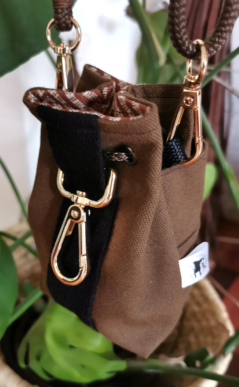 Puede incluir: Una bolsa de lona marr&oacute;n con una franja negra y herrajes dorados. La bolsa tiene un cierre de cord&oacute;n y un interior estampado. Se ve una peque&ntilde;a etiqueta blanca con una silueta negra.