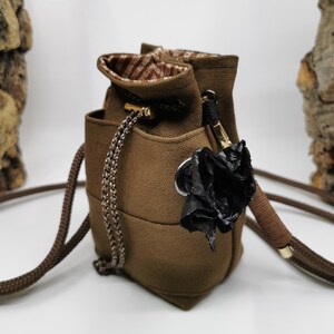 Puede incluir: Un bolso tipo cubo marr&oacute;n con cierre de cord&oacute;n y una correa larga y ajustable. El bolso presenta un forro interior estampado y una flor negra decorativa adjunta en el lateral. La correa mide aproximadamente 100 cm.