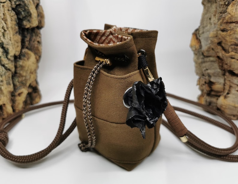 Puede incluir: Un bolso tipo cubo marr&oacute;n con cierre de cord&oacute;n y una correa larga y ajustable. El bolso presenta un forro interior estampado y una flor negra decorativa adjunta en el lateral. La correa mide aproximadamente 100 cm.
